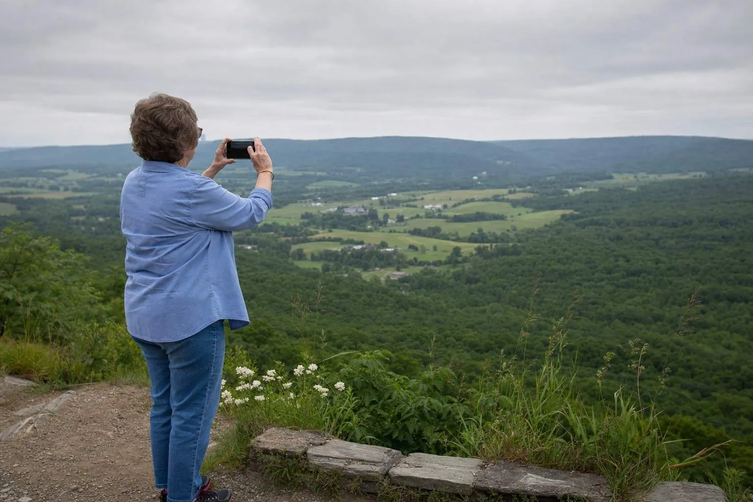 Side view of a person at a hillside overlook holding up a phone to a broad green valley