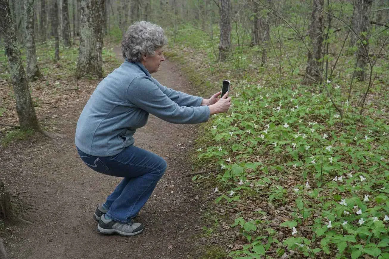 Side view of an everyday person crouching slightly on a wooded trail to frame a photo with their phone
