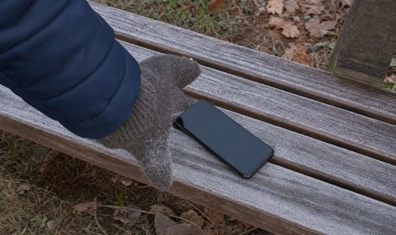 A smartphone lying on a frost covered bench with a gloved hand reaching for it