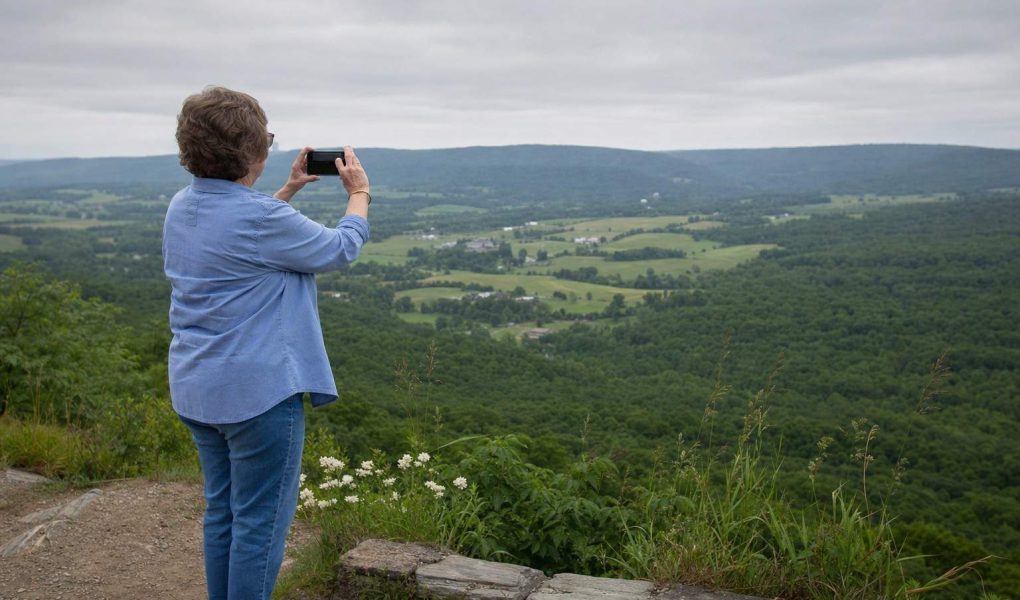 Side view of a person at a hillside overlook holding up a phone to a broad green valley