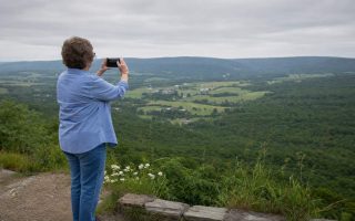 Side view of a person at a hillside overlook holding up a phone to a broad green valley