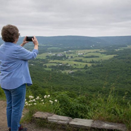 Side view of a person at a hillside overlook holding up a phone to a broad green valley