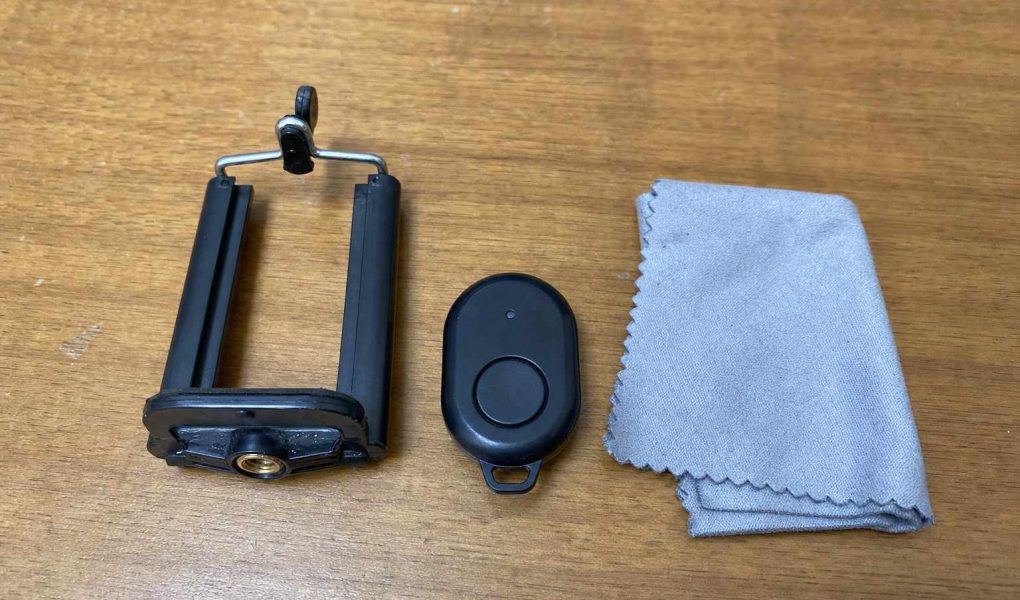 A small clip-on phone tripod mount, a tiny bluetooth remote, and a microfiber cloth laid on a plain wooden table