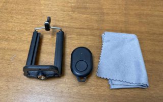 A small clip-on phone tripod mount, a tiny bluetooth remote, and a microfiber cloth laid on a plain wooden table