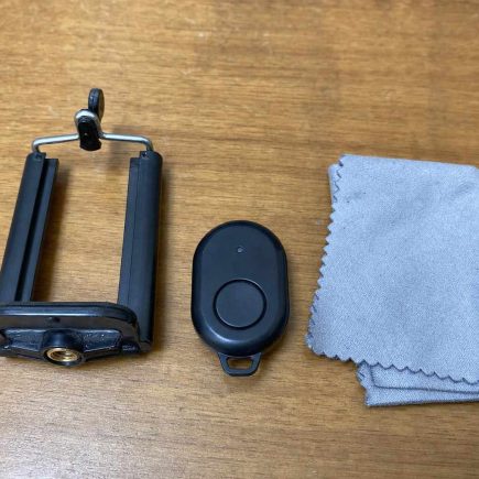 A small clip-on phone tripod mount, a tiny bluetooth remote, and a microfiber cloth laid on a plain wooden table