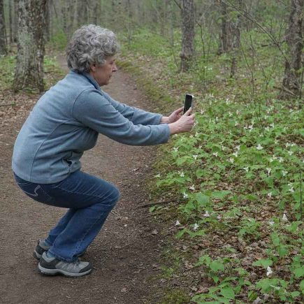 Side view of an everyday person crouching slightly on a wooded trail to frame a photo with their phone