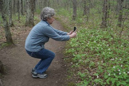 Side view of an everyday person crouching slightly on a wooded trail to frame a photo with their phone