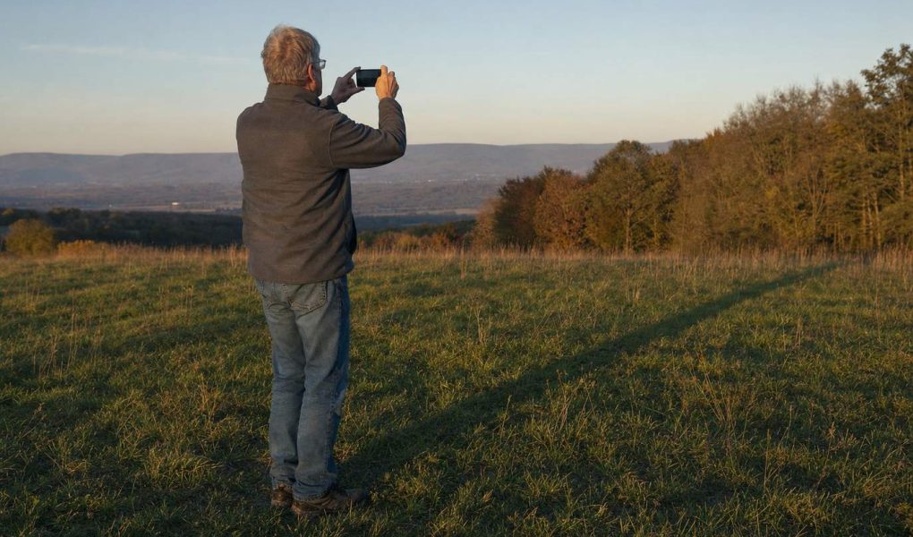 An older man holding up a phone to photograph long evening shadows across an open meadow
