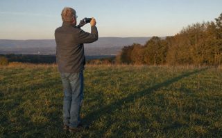 An older man holding up a phone to photograph long evening shadows across an open meadow