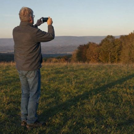 An older man holding up a phone to photograph long evening shadows across an open meadow
