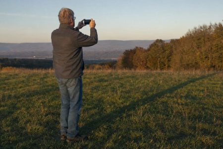 An older man holding up a phone to photograph long evening shadows across an open meadow