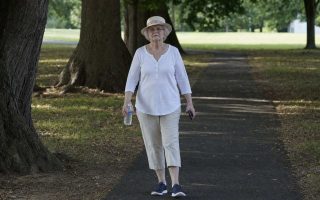An older woman in a sun hat walking a shaded park path early in the morning with a water bottle