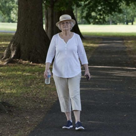 An older woman in a sun hat walking a shaded park path early in the morning with a water bottle