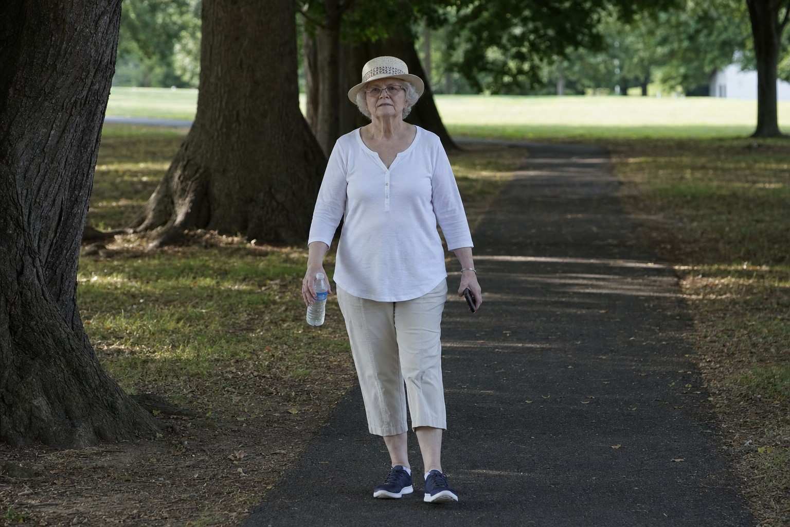 An older woman in a sun hat walking a shaded park path early in the morning with a water bottle