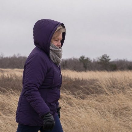 An older woman walking across an open windswept field in winter layers with her hood up