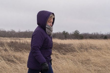 An older woman walking across an open windswept field in winter layers with her hood up