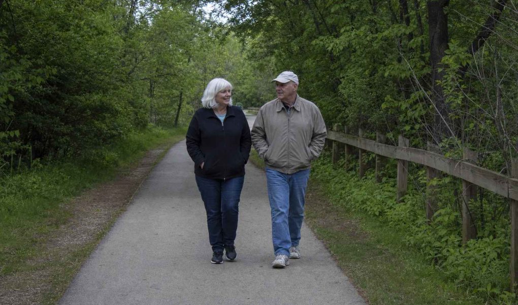 Two older people in plain casual clothes walking together at an easy pace along a tree-lined paved greenway path on an overcast day, hands relaxed in their jacket pockets