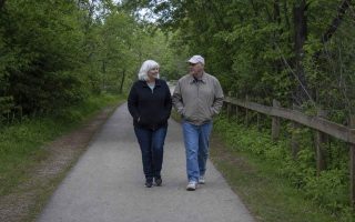 Two older people in plain casual clothes walking together at an easy pace along a tree-lined paved greenway path on an overcast day, hands relaxed in their jacket pockets
