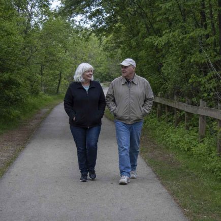 Two older people in plain casual clothes walking together at an easy pace along a tree-lined paved greenway path on an overcast day, hands relaxed in their jacket pockets