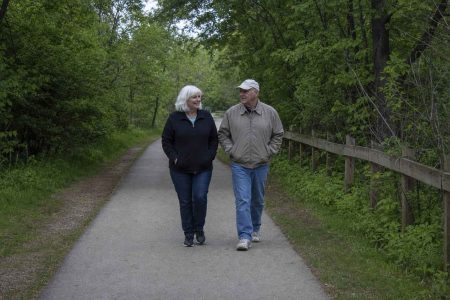 Two older people in plain casual clothes walking together at an easy pace along a tree-lined paved greenway path on an overcast day, hands relaxed in their jacket pockets