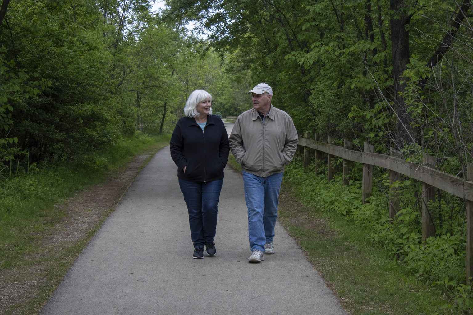 Two older people in plain casual clothes walking together at an easy pace along a tree-lined paved greenway path on an overcast day, hands relaxed in their jacket pockets