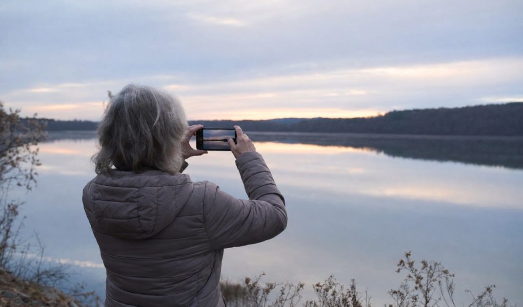 A person seen from behind in a warm jacket standing at a calm lakeshore at first light, holding up a phone to photograph the soft pre-sunrise sky