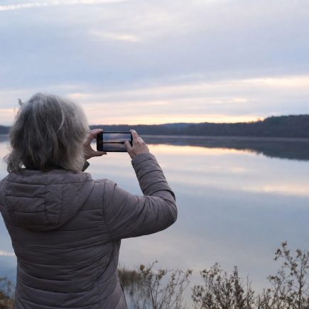 A person seen from behind in a warm jacket standing at a calm lakeshore at first light, holding up a phone to photograph the soft pre-sunrise sky