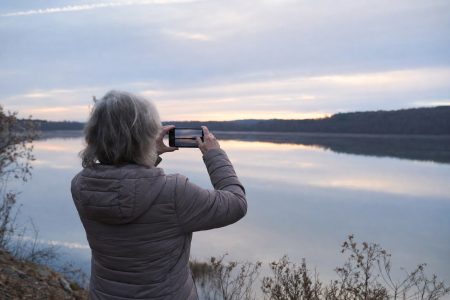 A person seen from behind in a warm jacket standing at a calm lakeshore at first light, holding up a phone to photograph the soft pre-sunrise sky