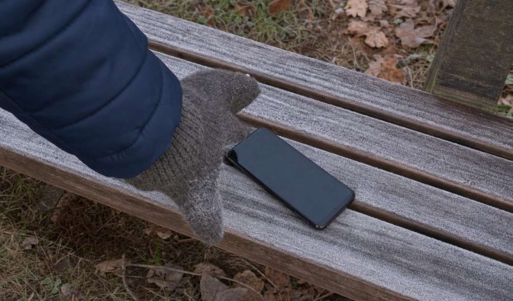 A smartphone lying on a frost covered bench with a gloved hand reaching for it