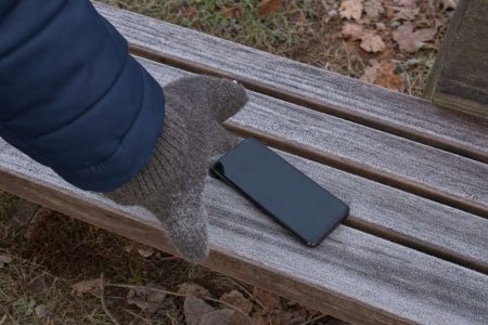 A smartphone lying on a frost covered bench with a gloved hand reaching for it