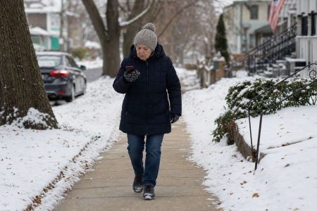 Front view of an older person in winter layers walking carefully on a snowy sidewalk