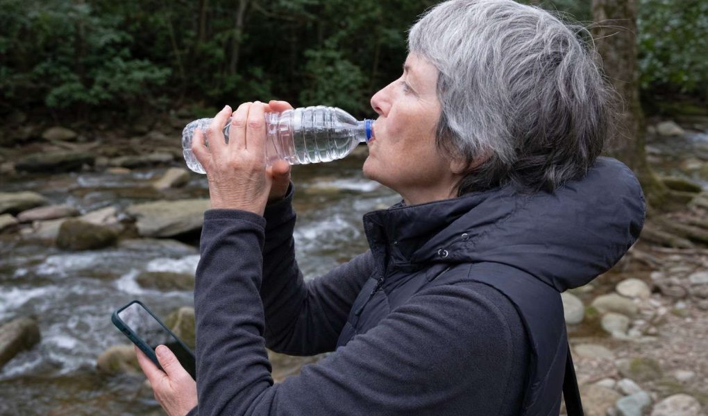 Side close view of an older walker drinking from a water bottle on a trail rest stop near a creek