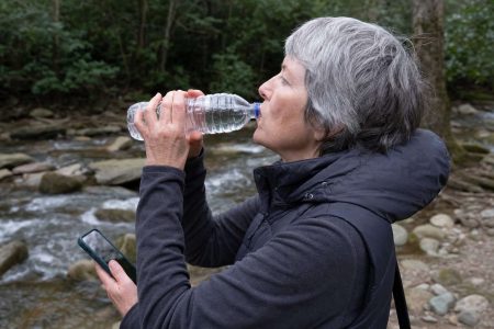 Side close view of an older walker drinking from a water bottle on a trail rest stop near a creek